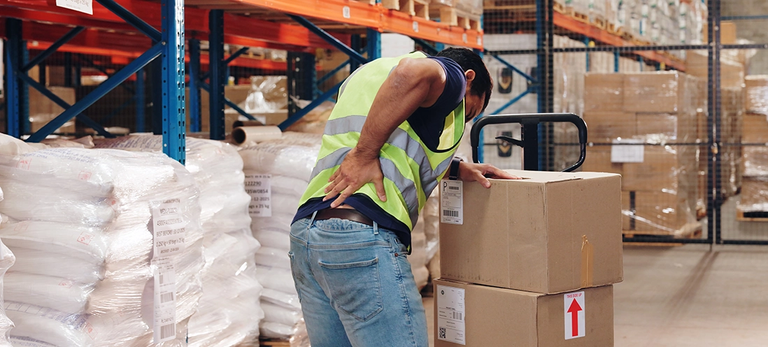 A person in a safety vest holds their lower back in pain in a warehouse.