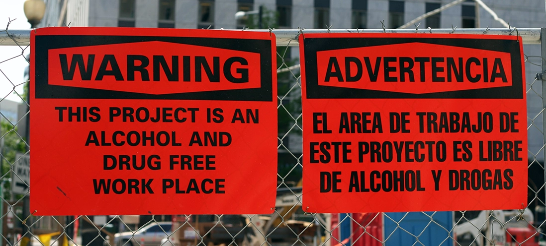 Two signs on a fence: one in English and one in Spanish, both stating the workplace is alcohol and drug-free.