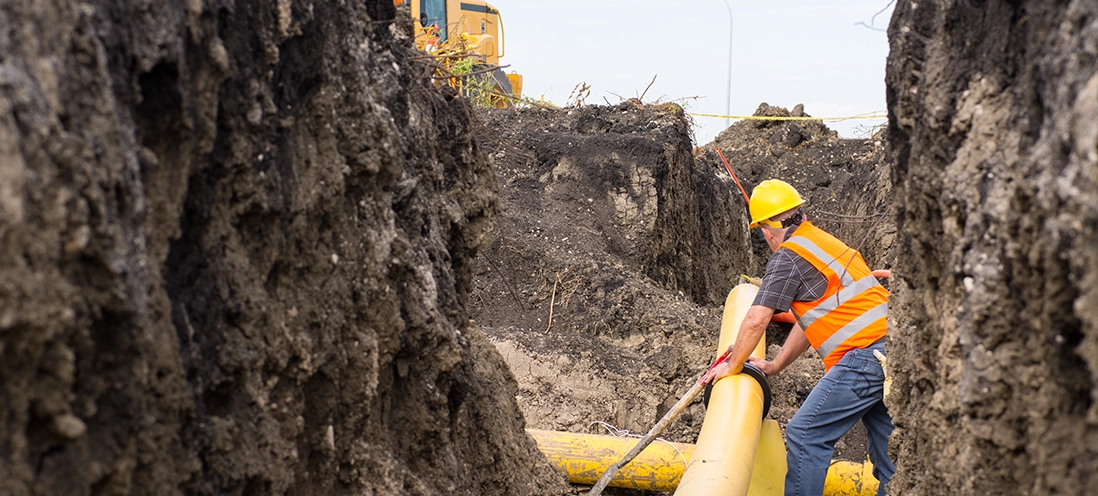 A construction worker in a safety vest and helmet handles a large yellow pipe in a dirt trench.