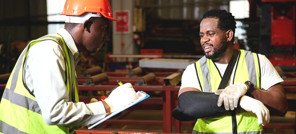 A supervisor and injured employee converse on a job site. Supervisor takes notes on a clipboard.